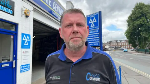 Mark looking into the camera with a straight face. He's wearing a black polo shirt with royal blue collars and shoulders, with a blue and white logo on the left chest reading 'Chase car care centre'. He's stood in front of his garage, which is a single-storey whitewashed building with a blue sign above it - in white letters his company name is written.