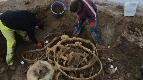 Durham University Two people standing in a big muddy hole, bending down to examine large wheels covered in rust and mud 