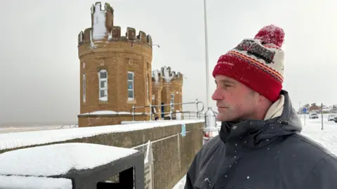 Kevin Shoesmith/BBC A man in his 40s, wearing a winter coat and a red bobble hat, looks out to sea as snow falls. The sky is grey and the sea rough.