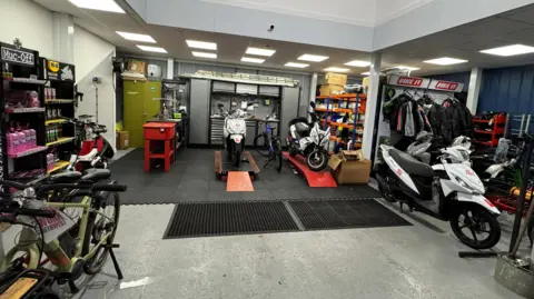 A picture of the warehouse. It has a number of bikes with repair stations. There is items around the warehouse, with motorbikes also in the distance.