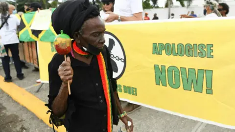 A protester wearing a black shirt and hat walks past a sign reading "apologise now", outside the entrance of the British High Commission during the visit of the Duke and Duchess of Cambridge in Kingston, Jamaica on 22 March, 2022