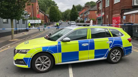 A police car parked across a road, with police tape stretched across it, between rows of houses and shops.
