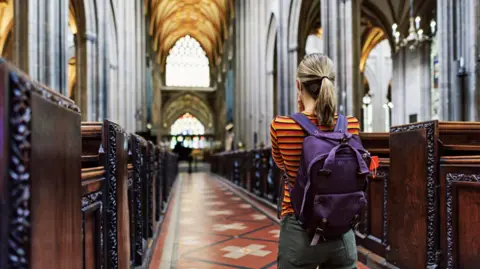 Imgorthand/Getty The back view of a young woman looking down the aisle of a church with wooden pews either side of her. She has a dark blonde ponytail and is wearing grey-green trousers, an orange, black and red striped top and has a purple rucksack.
