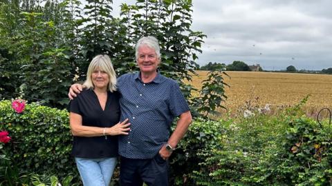 Bill Doran and his wife standing in a garden with a view over the Suffolk countryside behind them
