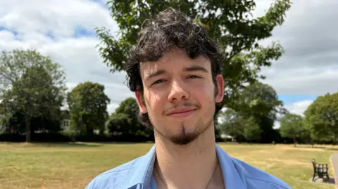 Finnley smiles at the camera. He has short curly dark hair and a short goatee. He wears a blue shirt and stands in a park. 