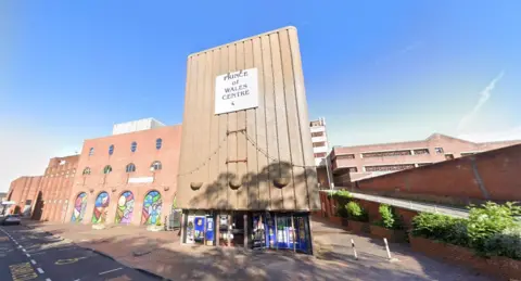 Google A large building with a ramp at the side of it. There is a sign which reads Prince of Wales Centre above a set of windows and glass doors. A section of the building set back from the entrance has a number of archways which each contain colourful murals.