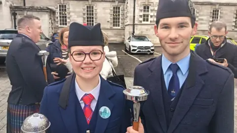 Georgia Turkington is wearing black-framed glasses and a navy uniform and red tie.
Louis Anderson has short dark hair and is also wearing a navy uniform with a navy tie and light blue shirt   