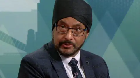 A man in a navy suit and black turban. He is sitting on a sofa and looking to the left. He is wearing a white shirt and floral tie, as well as rectangular glasses.