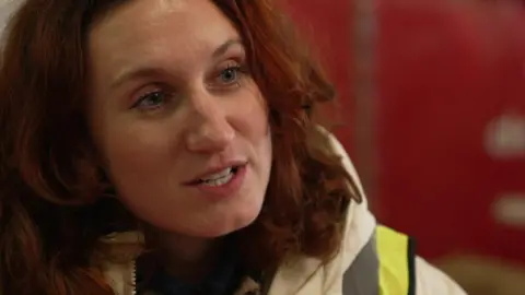 A close up of Bessie Carter's face. She is wearing a white coat and a high vis tabard and has long dark red hair. 