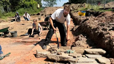 Oxford Cotswold Archaeology A man wearing a white t-shirt and black trousers digging at the site of the former Tudor mansion site