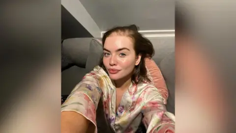 Olivia McVeigh Olivia McVeigh taking a selfie. She is wearing a floral dressing gown and is sitting on a sofa. 