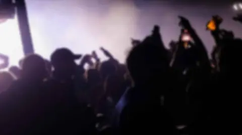 Blurred image of people dancing holding their phones in the air at a music festival. In the background is white smoke and purple lights.