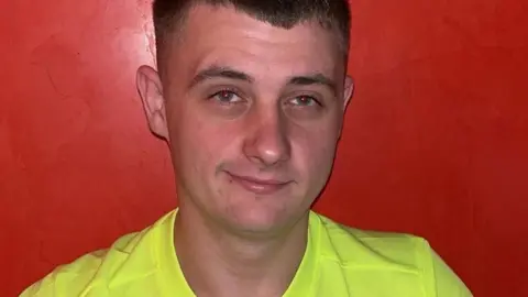 Fraser Stewart is half smiling at the camera. He has short, brown hair, shaved at the sides and is wearing a luminous yellow T-shirt. He is standing against a red wall.