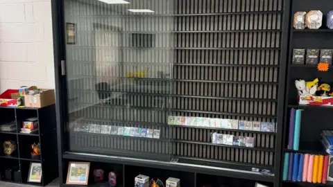 Juliano Scibelli A black standing glass shelf standing empty with some cards placed on a rack at the bottom.