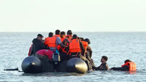 A group of people, many wearing life jackets, are on a small boat off the coast, with two others trying to climb aboard.