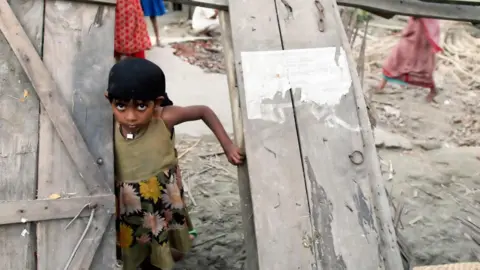 Getty Images A young girl with dark hair and big eyes looks out through the doorway of a wooden building that has been shattered by the storm. She looks sad and anxious.