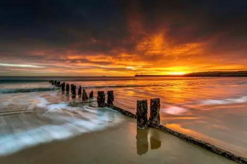 Malcolm Wilson Sunrise on Aberdeen beach, showing wooden spines going into the sea, and an orange glow in the background.