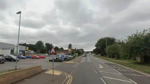 A road with a Lidl supermarket on one side and a row of fences and trees at the other.