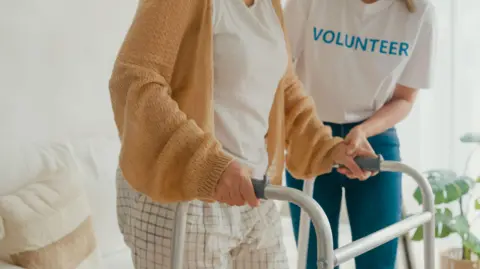 Closeup of young female volunteer teach old woman to walk use walker take care hold hand elderly patient in living room at home. The volunteer has "volunteer" written across her t-shirt.