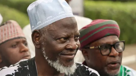 AFP via Getty Images Sani Abdulkadir with a white beard and a pale blue robe with black embroidery and matching traditional pale blue hat smiles as he arrives at court on Wednesday 22 April 2026.