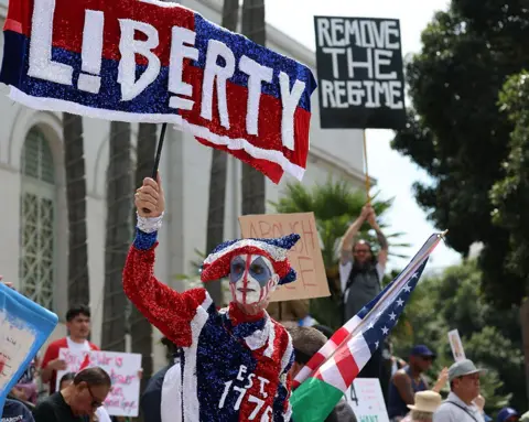 Getty Images A person is seen wearing a red, white and blue sparkly costume with their face painted. The front of costume reads 1776. Others behind this person can be seen holding up protest signs against Trump. 
