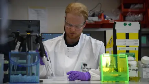 A lab technician wearing a white jacket, purple gloves and protective glasses while preparing samples. He has fair hair and is sitting at a desk.