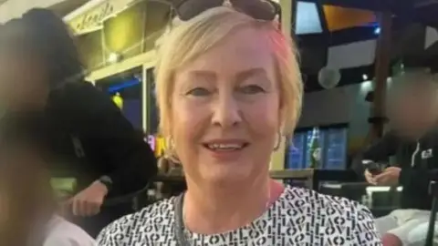 A woman in her fifties with blonde hair. She is wearing sunglasses on her head and appears to be sat in a bar. People behind her have their faces blurred.