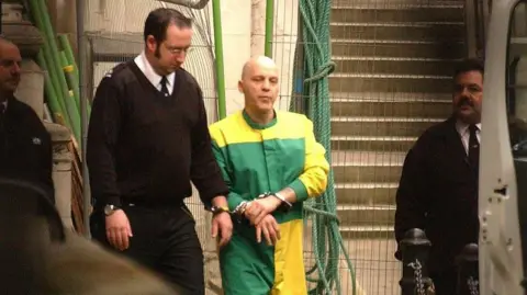 PA Media A shaven-headed Michael Stone in 2005. He is handcuffed to a prison officer and is wearing a green and yellow jump suit. He appears to be being led towards a van.