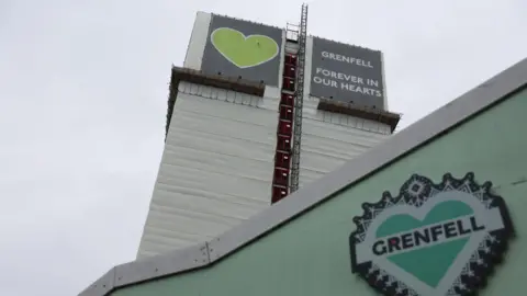 Reuters The Greenfell Tower is seen on from a perspective looking up with a green memorial wall seen in the foreground. It features a heart with "Grenfell" written across it. 