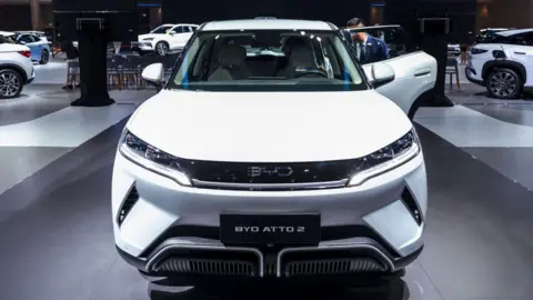 A white BYD ATTO 2 made by Chinese electric vehicle manufacturer BYD at the 2025 Bangkok International Motor Show at the Impact Arena convention venue in Bangkok. A man in a mid-blue jacket, white shirt and blue-striped tie is looking into the left hand back door. There are other cars and people in the background.