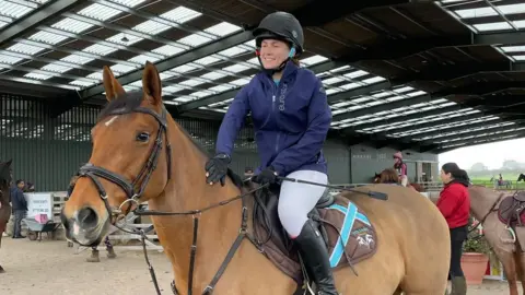 Millie Tingey Millie Tingey sits on her bay horse in a riding school, surrounded by other people on horses and spectators, in English horse riding attire. She is smiling as she pats her horse, Baby, on the neck.