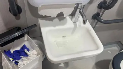 Sarah Cadwallader Blocked small white sink with water level risen up to halfway and a tissue paper floating in it. A wastepaper bin is next to it.