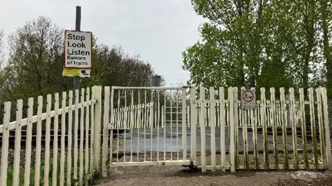 A level crossing, pictured from behind its pale-coloured gate and fence. A 'Stop, Look, Listen' sign is visible.