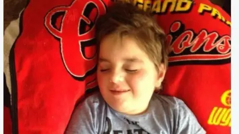 Paul Woodward Kalel is lying on a bed with his eyes closed. He is wearing a grey Beatles T-shirt and is lying on a red Grand Prix blanket. 