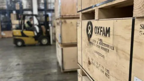 A wooden crate with the Oxfam logo printed on it. Behind, out of focus, is a man driving a forklift truck.