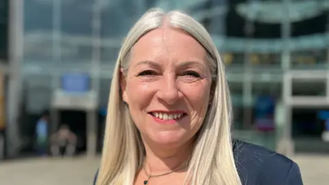 A head and shoulders shot of Kay Mason Billig in front of The Forum, Norwich. She has long blonde hair and is wearing a navy blue jacket and top.