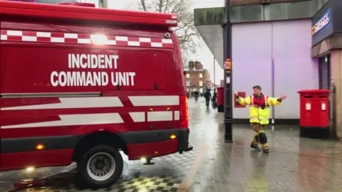 A red Incident Command Unit van can be seen parking in the high street outside a Greggs store. An emergency worker in high-vis can be seen directing it. 