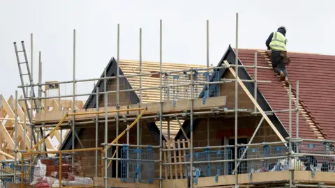 A construction site showing a house in progress, with scaffolding surrounding the structure and a worker in a high-visibility vest installing roof tiles on the sloped roof. Wooden beams and other construction materials are visible in the foreground and background.