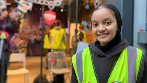 Jon Wright/BBC A girl wearing a high-vis jacket stands in front of a glass shop window with chains and keys painted on.