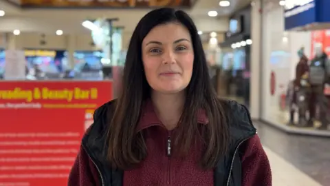 A woman with long dark brown hair, looking at the camera. She has a red fleece and a black coat on.