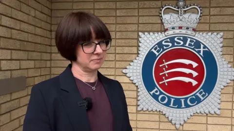 Stuart Woodward/BBC A woman with dark brown hair looks at the camera. She is wearing glasses and a black blazer with a top beneath. There is an Essex Police sign behind her. She has a serious look on her face.