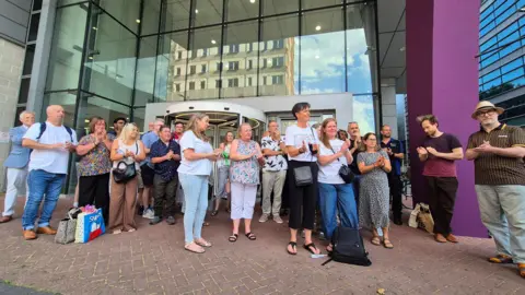 Stuart Woodward/BBC Several members of staff gathered outside the Gateway building of the University of Essex's Southend campus and applaud. The building has a large glass front with a purple pillar on the right hand side.