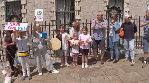 Protestors gather in Torquay ahead of council decision. There are both male and female protestors present. They are banging instruments and holding up banners. Young children are in attendance.
