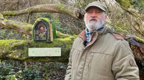 A fairy door with a black door is seen, John Rowe in the foreground. He has a cap on and and brown jacket and has a grey beard.