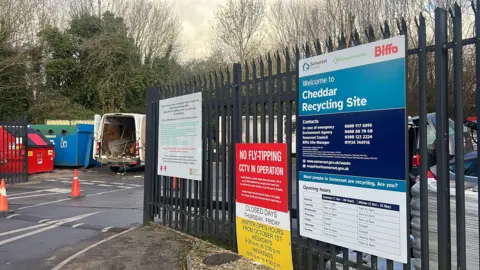 The gated entrance to the recycling centre. It features three signs on the fence - two with rules and warnings, the other with opening hours. In the background through the gate you can see a van with its doors open with scrap metal inside.