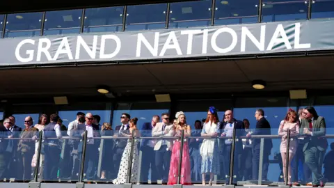Smartly-dressed spectators look out from a balcony under a large banner saying "Grand National"