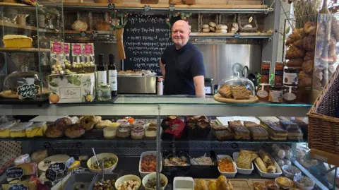Hayley Coyle/BBC A shot of the interior of GG's Artisan Bakery and Deli in Holmfirth with the owner stood behind a deli counter containing pastries, cakes, cheese and olives.