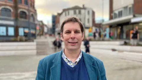 A man stood on a high street looking at the camera. The background is blurred but shows an array of shops. He is wearing a light blue suit jacket and a darker blue sweater vest. 