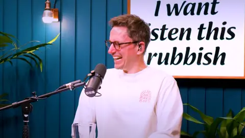 John Robins sits in front of a microphone, he laughs as he looks at his co-host. 