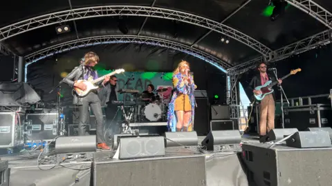 A female signer wearing a flowing, purple, blue and yellow dress leads a four-piece band, including two guitarists and a drummer, on a stage with black tarpaulin-style walls and roof at Humber Street Sesh in Hull